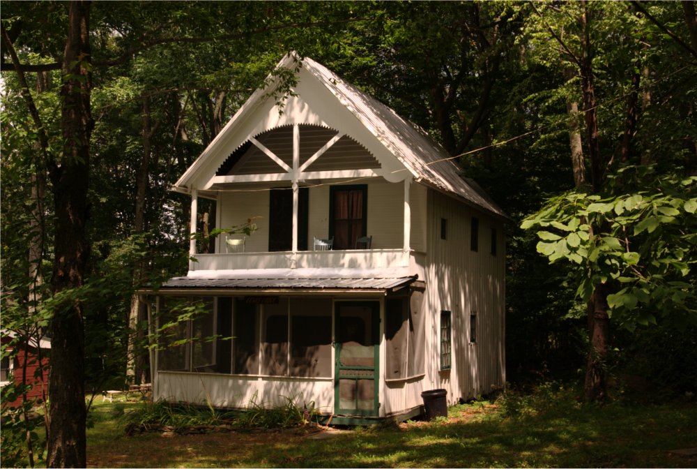 A two-storey white cottage with a gabled porch and decorative trim, set among the trees.